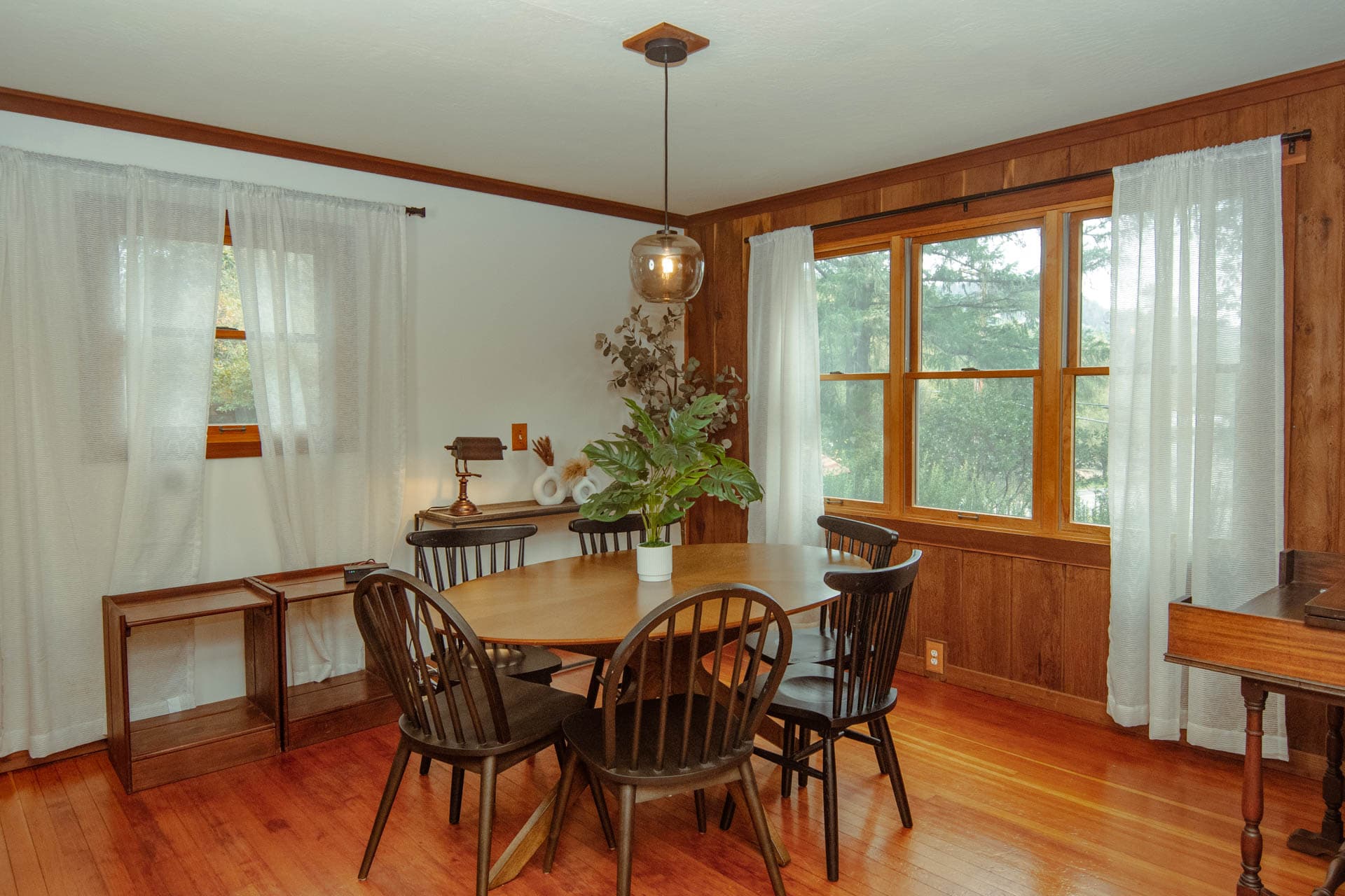 A cozy dining area featuring a wooden table surrounded by black chairs, with large windows and light curtains.