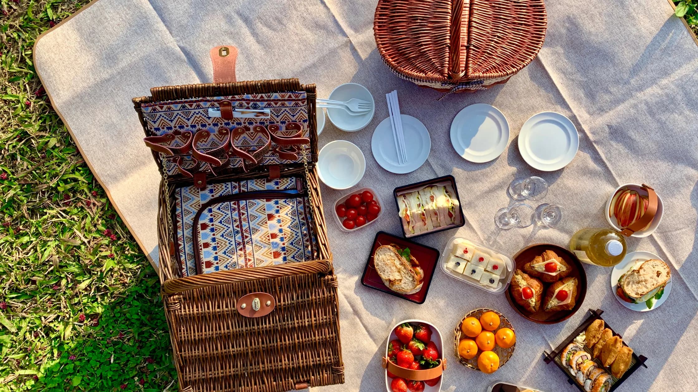 A picnic spread with a wicker basket, plates, glasses, and an array of food items on a blanket in a grassy area.