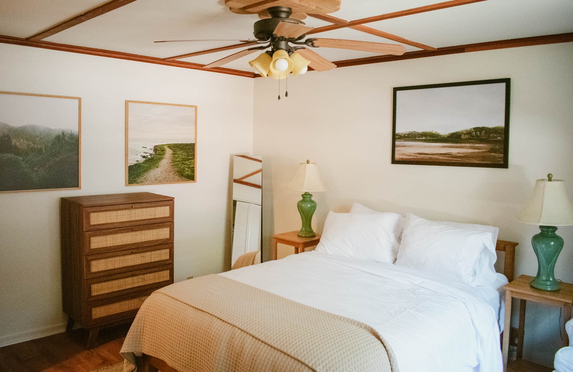 A cozy bedroom featuring a bed, nightstands with green lamps, a wooden dresser, and framed landscape pictures on the wall.