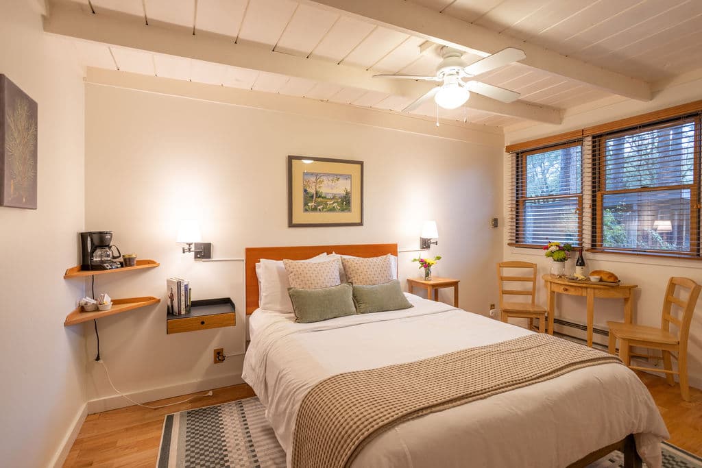 A bedroom with a queen bed with white linens and a tan throw, two nightstands with lamps, a small table with chairs, artwork on the wall, and a wooden beam ceiling.