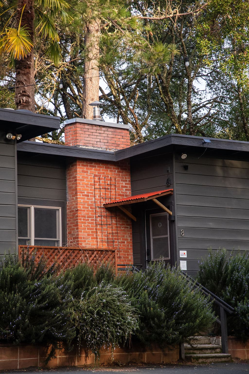 A modern gray house with a brick chimney and an overhanging red roof, surrounded by lush greenery.