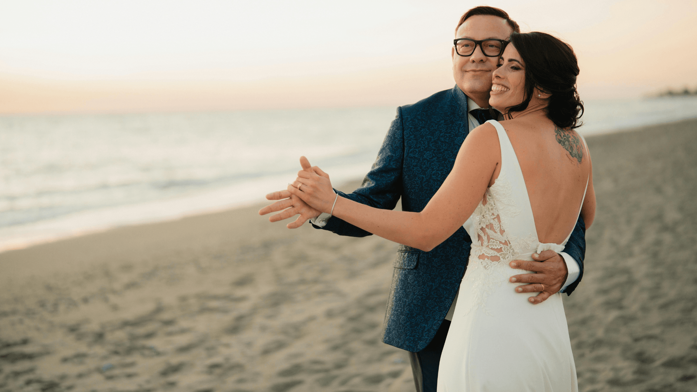 A couple dances together on a beach at sunset, smiling and enjoying their moment.