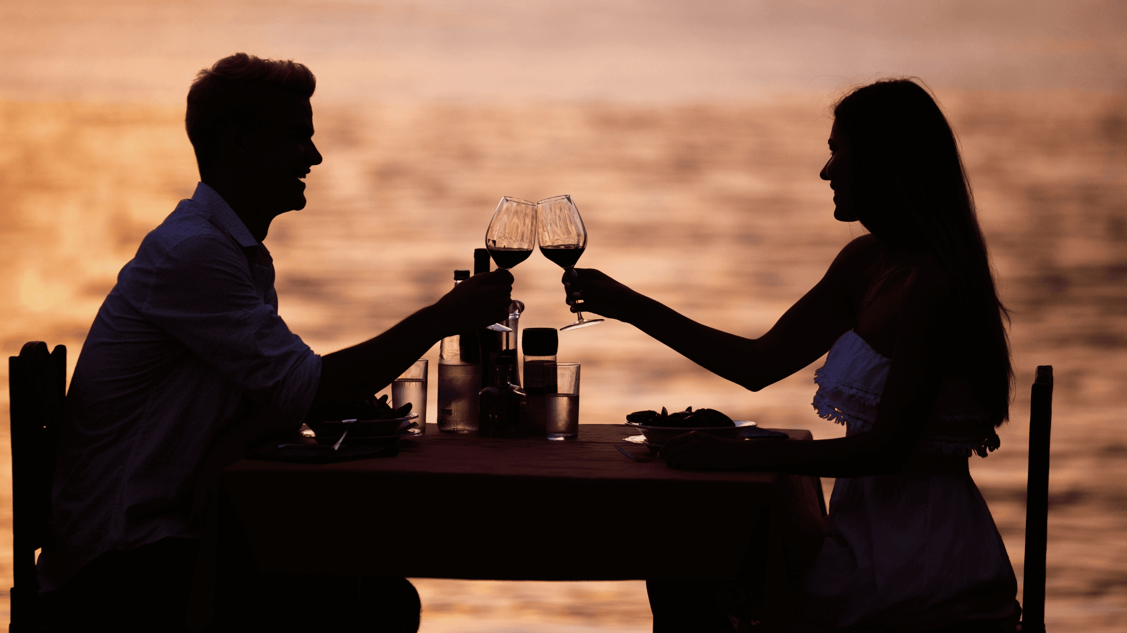 A couple clinks wine glasses at sunset, silhouetted against the water.