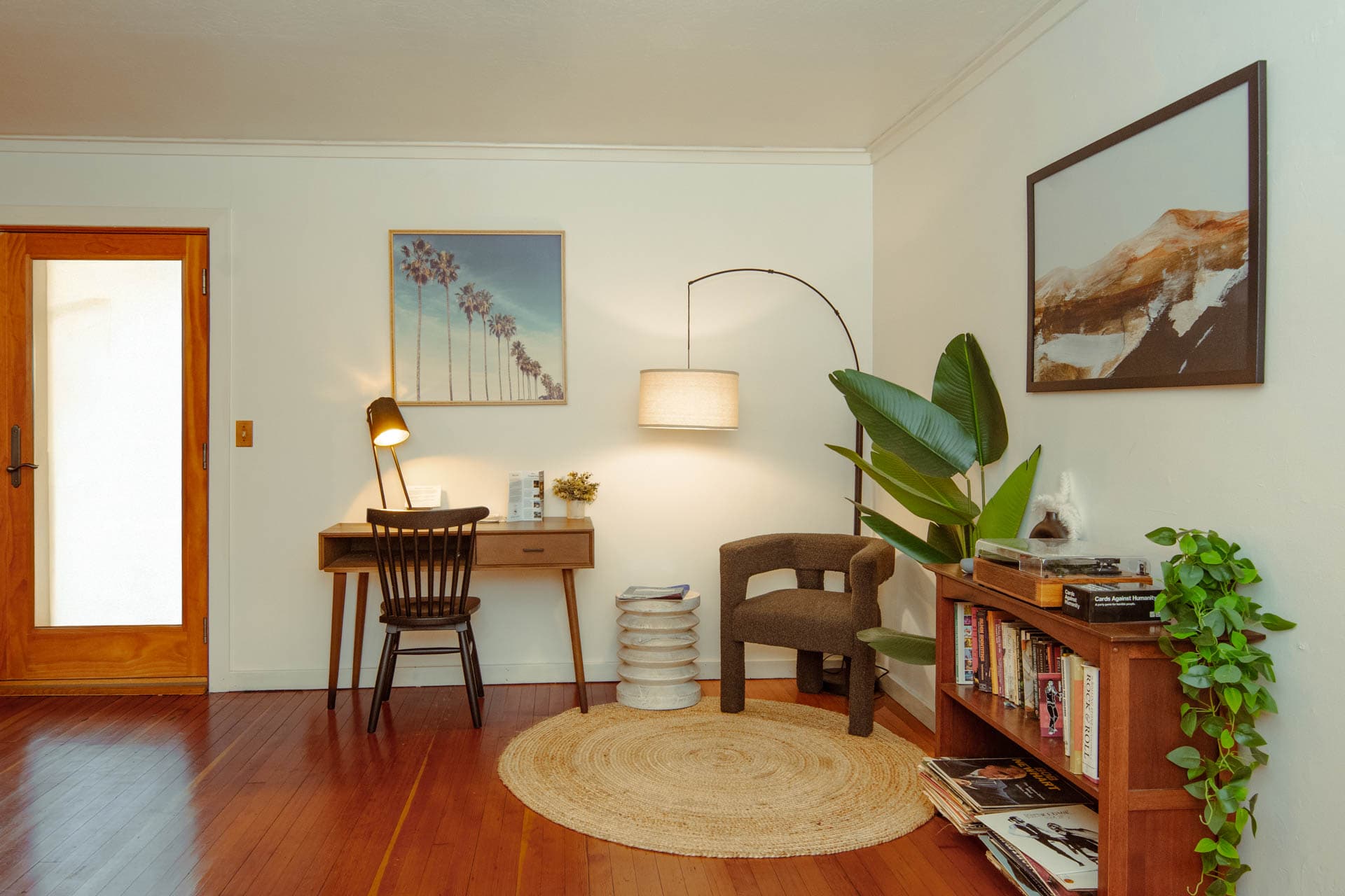 A cozy living room featuring a desk with a lamp, a comfortable chair, potted plants, and artwork on the walls.